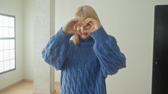 Woman forms heart with hands and smiles toward camera, wearing chunky blue turtleneck sweater and bangs in bright studio with grid window; love joy.