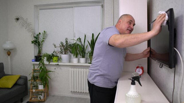 An elderly man is meticulously cleaning a television screen in a comfortable living room adorned with potted plants. He uses a cloth and spray to ensure clarity and shine.