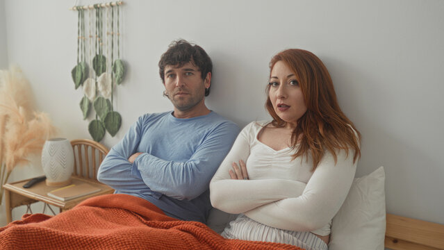 Woman and man sit on bed with crossed arms under orange blanket in studio, woman looks away while man closes eyes; relationship tension.
