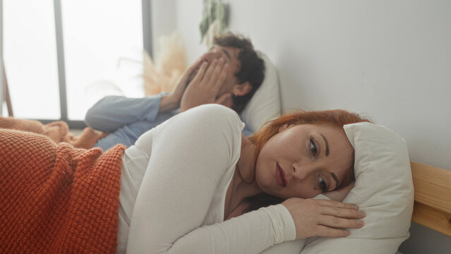 Woman lying on bed under orange blanket with hand near face while man leans over and touches her shoulder in bedroom; tension.