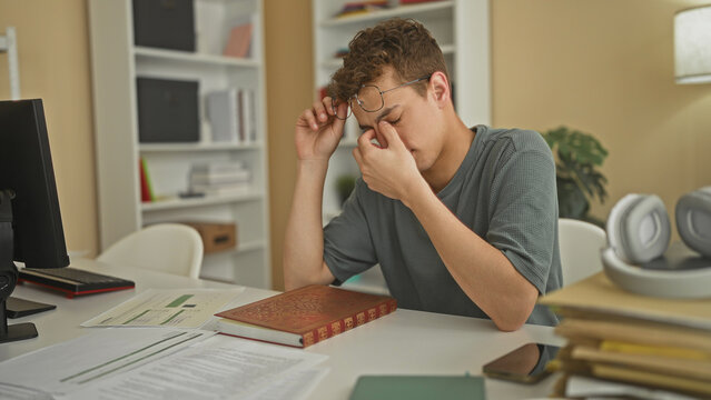 Young man flips ornate red book page, adjusts glasses and reaches for pen at a desk in a building study; concentration.