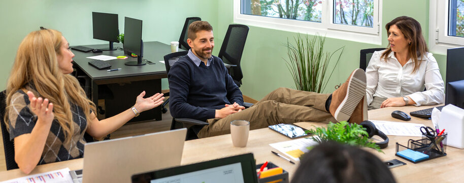 Smiling man rests feet on shared desk while female colleagues react with questioning expressions and raised palms. Office etiquette concept highlighting disrespect, tension, inappropriate behavior.