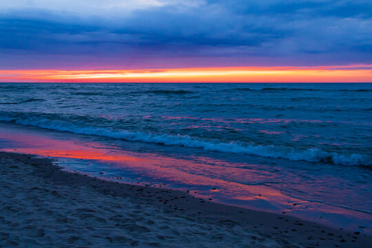 Pink Sunset Reflection Across Dark Ocean and Sandy Beach. A narrow pink and orange sunset glows along the horizon above a deep blue ocean. The warm colors are reflected across the wet sandy beach 