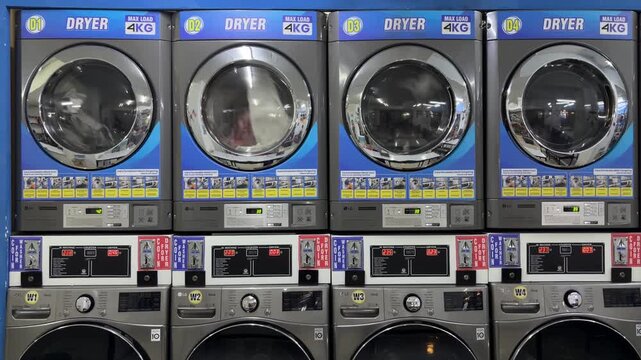 A row of modern coin-operated dryers in a laundromat. Each dryer has a circular glass door and digital control panel. The setting is clean and well-lit.