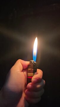 a portable lighter igniting and burning in pitch black darkness. A steady, bright yellow and blue flame illuminates a person's thumb in the foreground. Cinematic low light concept of fire, heat