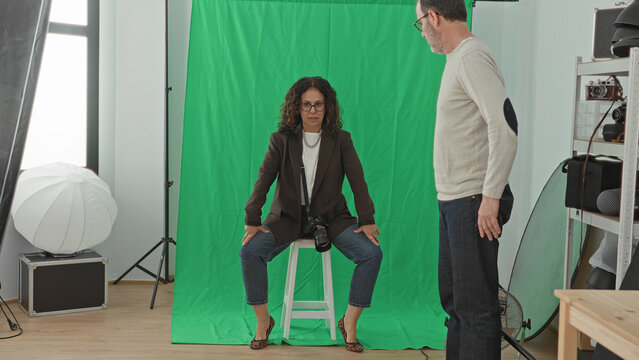 Woman photographer with camera directs seated man model on stool against green screen backdrop in studio; focused collaboration.