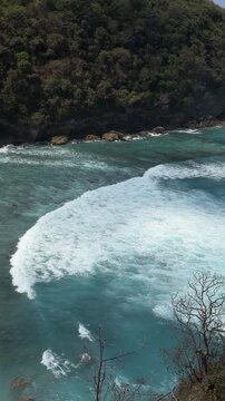 A scenic view of a turquoise ocean with white waves crashing against the shore. Lush green hills rise in the background, creating a tranquil coastal landscape.