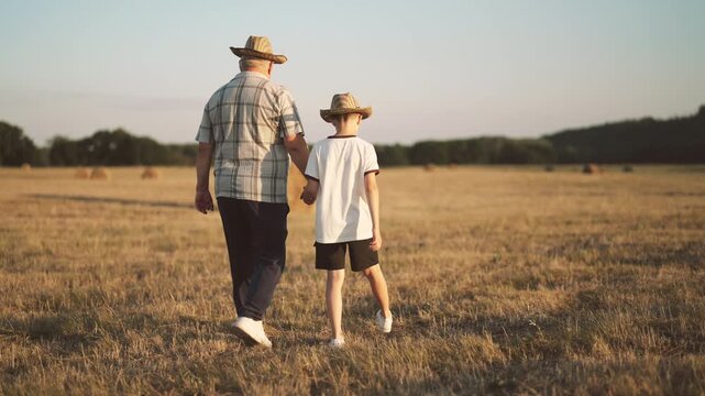 Curious child and his grandfather walking in farmland, back view, eco-farming. Harvest season in countryside, grandpa and grandson strolling in wheat fields, admiring haystacks in ranch, straw hats