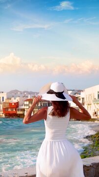 A tourist woman in a white summer dress looks at the famous district of Little Venice in Mykonos, Cyclades island, Greece, during sunset