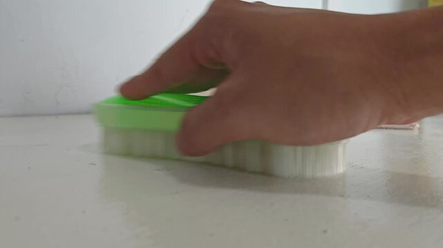 a person's hand using a green plastic bristle brush to scrub and clean a white surface. Concept of household chores, spring cleaning, sanitation, hygiene, and domestic maintenance
