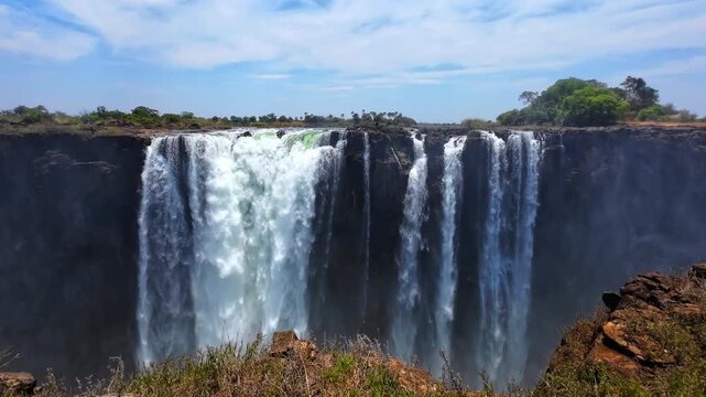 Broad and narrow curtains of water spill over the dark cliff at Victoria Falls, with mist rising from the gorge and trees beyond the riverbank.