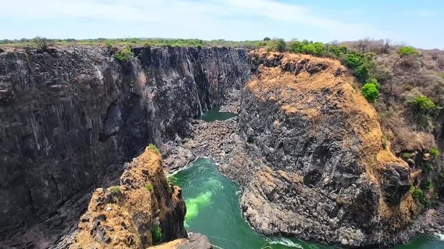 A dry, grass-covered rock formation rises above the Zambezi River as green water curves through a steep gorge below Victoria Falls.