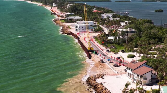 Yellow crane and crews reinforce a beachfront seawall along a narrow coastal road beside bright green ocean water.