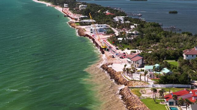 Construction crews build a new coastal seawall beside upscale waterfront homes along a green tropical shoreline.
