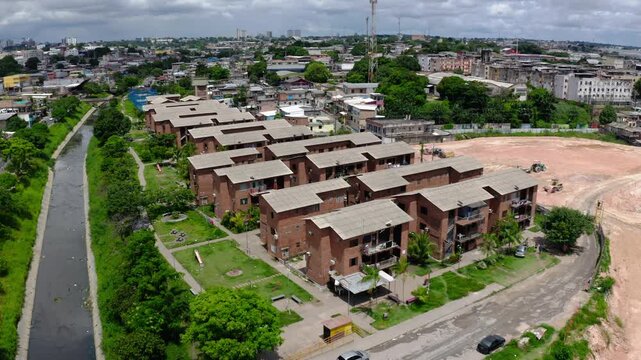 Static aerial drone view of rows of brick social housing blocks of a conjunto habitacional beside the canalised igarape in Manaus, lawns, palms, parked cars, graded lot, city skyline, sunny, Brazil.
