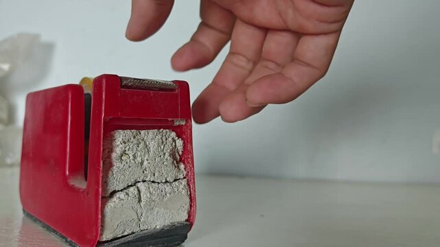a person's hand pulling and tearing clear adhesive tape from a heavy-duty red desktop tape dispenser. Concept of office work, school supplies, packaging, art and crafts, and everyday desk organization