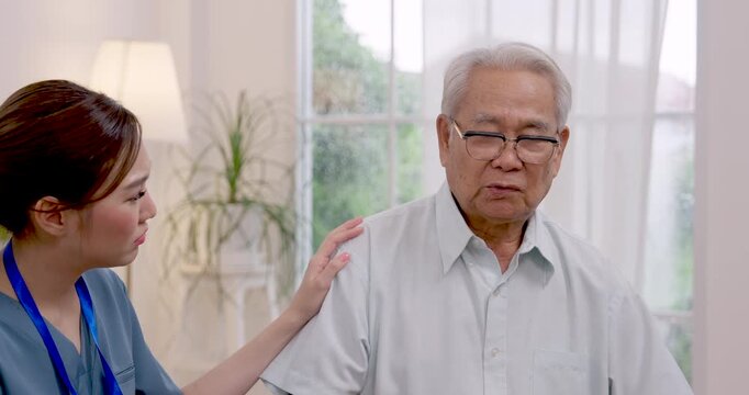 Asian nurse listens to senior Asian man, gently touches shoulder and offers reassurance while resident discusses health concerns and rehabilitation needs inside modern assisted living community.