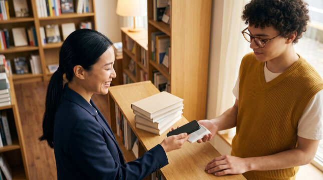 Woman tapping smartphone on card reader at bookstore checkout illustrating contactless payment technology and retail point of sale concepts.