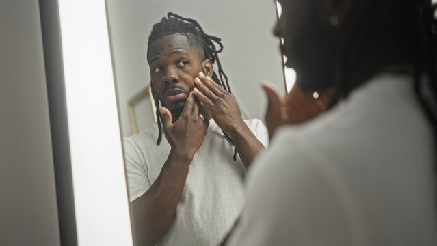 Young man touching cheek and examining skin in a lighted mirror at studio vanity while adjusting rings on fingers; self care confidence.