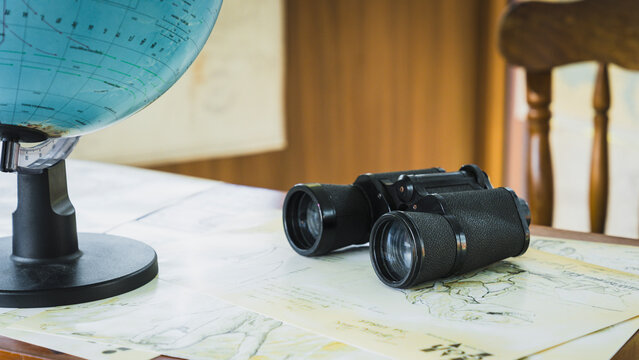 Travel exploration and navigation concept, vintage binoculars resting on antique world map beside desktop globe on wooden desk, symbolizing discovery, geography, wanderlust, and adventure.