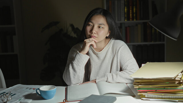 Woman with hand on chin studying at desk with open notebook, blue cup, lamp and tall stack of folders, bookshelf visible in a studio; contemplation.