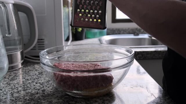 A person grates an ingredient on a metal grater into a clear glass bowl with ground meat on a kitchen countertop.