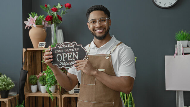 Man smiling and holding open sign with hand in florist shop building among potted plants and wooden crates; welcoming service.