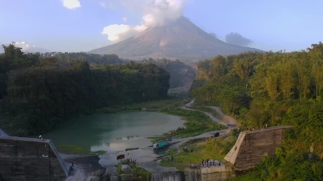 Stunning aerial drone shot capturing the iconic Mount Merapi (Gunung Merapi) active volcano towering over a scenic landscape in Magelang, Central Java, Indonesia. The video features a large check dam 