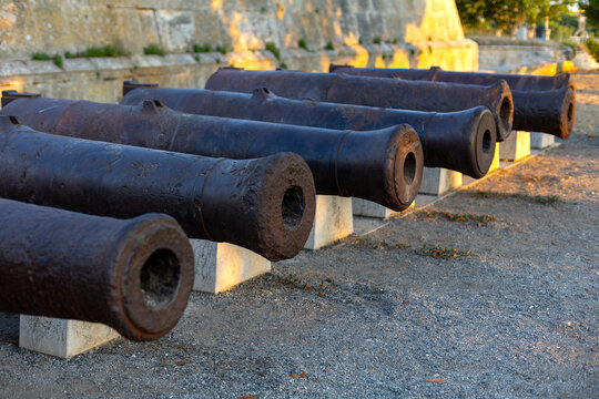 Historic Iron Cannons Lined Up at Stone Fortress. Rows of old iron cannons displayed beside a stone fortress wall in warm evening light.