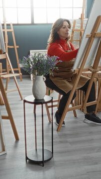Woman in apron painting on a canvas at an art studio, brush touching canvas as she sits at an easel; serene focus.