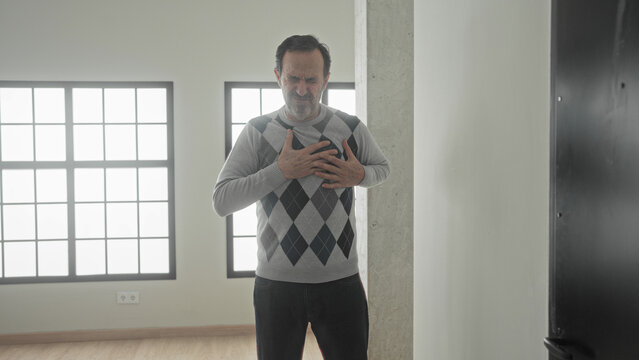 Middle aged man clutch chest with one hand, leaning on wall near window and pillar, grimacing and showing heartattack in building; pain emergency.
