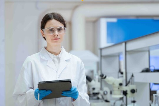 Female scientist wearing safety glasses and lab coat, smiling while analyzing data on a tablet in a modern research laboratory