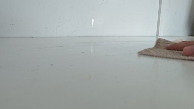a person's hand using a brown microfiber cloth to carefully wipe and clean a white table or counter surface indoors.