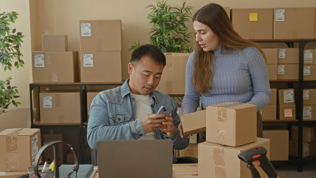 Man points smartphone at cardboard box while woman hands over a parcel and gestures toward stacked boxes in building; teamwork focused.