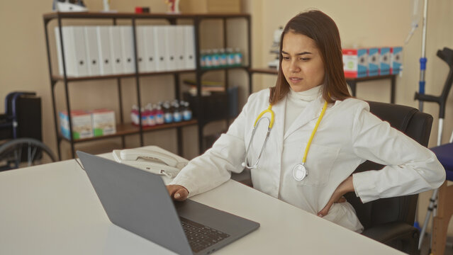 Woman doctor with yellow stethoscope sitting at desk typing on laptop while holding lower back in building medical clinic; backache fatigue.