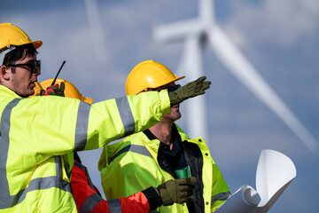 Fototapeta premium Two wind technicians with drawings shout observations over the rotor noise directly beneath a turning wind turbine in bright breezy daylight on a working wind farm in rural Thailand.