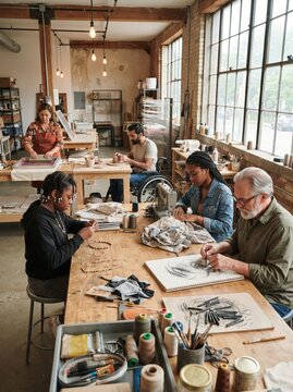 A diverse group of people work on different craft projects together in a spacious studio, including sewing, pottery, sketching, and beadwork at shared wooden tables.