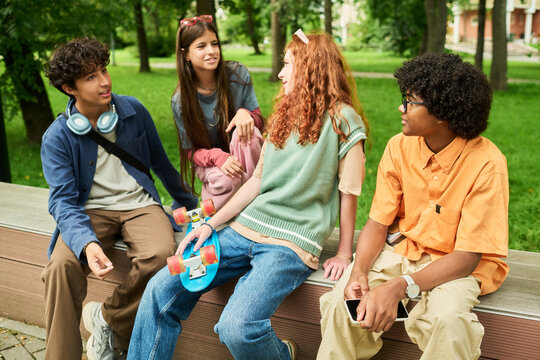 Multiethnic teenagers talking and relaxing on bench in park with skateboard and smartphone. Lifestyle image suitable for youth marketing campaigns and social media advertising