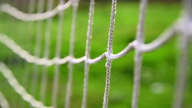 Soccer goal net fills the frame, woven ropes and knots forming a repeating texture against a soft, out of focus green grass playing field in daylight, empty stadium feel
