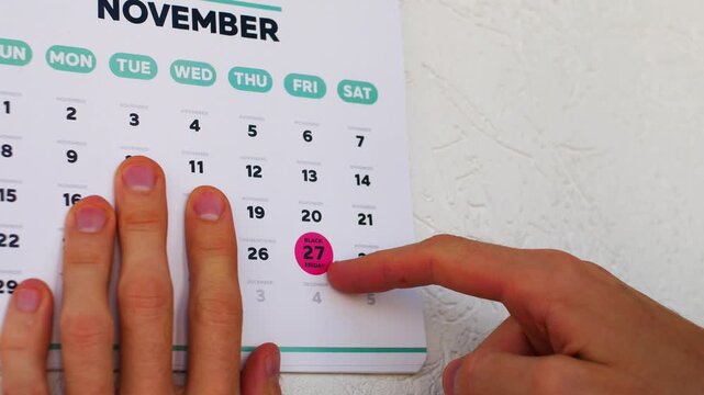 A man's finger pointing to the marked Black Friday date on a November 2026 American layout wall calendar sheet. Autumn shopping season consumerism and big sales discount concept