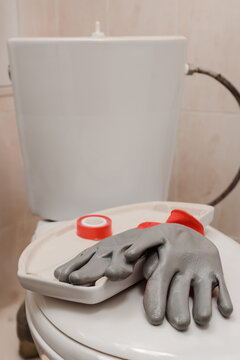 Close-up of toilet cistern with lid removed, exposed water hose and internal fittings visible, grey rubber gloves resting on lid, red tape roll nearby, showing bathroom repair work in progress