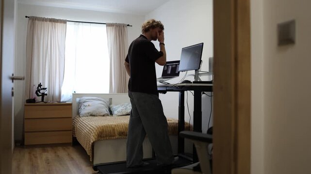 Young man walks on a treadmill while adjusting his standing desk setup combining light exercise with computer work for a healthier productive home office routine daily