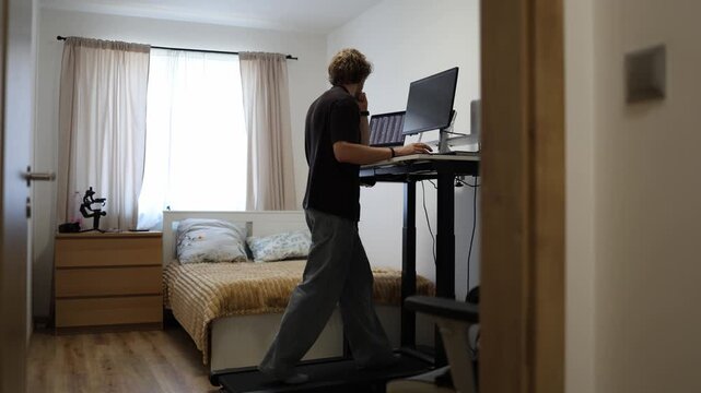 Young man walks on a treadmill while working at a standing desk with computer screens combining remote work fitness productivity and an active home office routine indoors