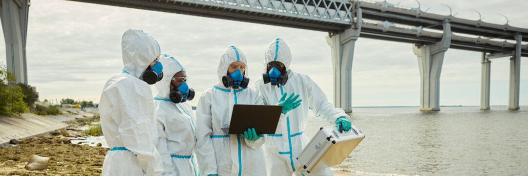 Header of multiethnic group of scientists wearing hazmat suits analyzing data on laptop outdoors near polluted water. Visual represents ecological research, environmental safety inspection