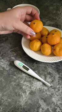 The woman is taking tangerines from a plate; there is a thermometer on the table. The concept of consuming vitamin C during a cold. ARVI, ARD, virus.