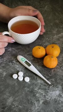 Woman drinking hot tea while sick with a cold, with tablets and a thermometer on the table, illustrating treatment of respiratory illness, seasonal flu and viral infection.