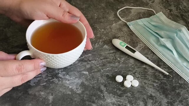 Woman drinking hot tea while sick with a cold, with tablets and a thermometer on the table, illustrating treatment of respiratory illness, seasonal flu and viral infection.