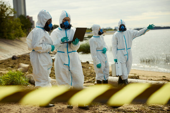 Diverse team of ecologist scientists in protective suits using laptop on polluted riverbank. Scene represents contamination inspection suitable for ecological research reports