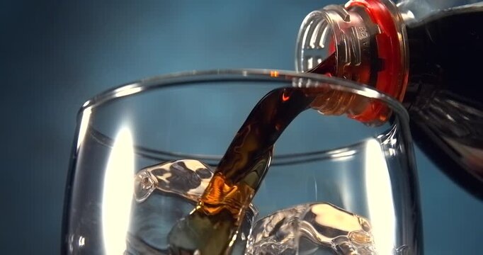Cola type sweet fizzy drink pouring into glass with ice cubes in slow motion on blue background