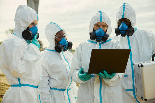 Diverse group of scientists in hazmat suits and respirators examining data on laptop outdoors. Perfect for environmental research, biohazard safety and epidemic control campaigns